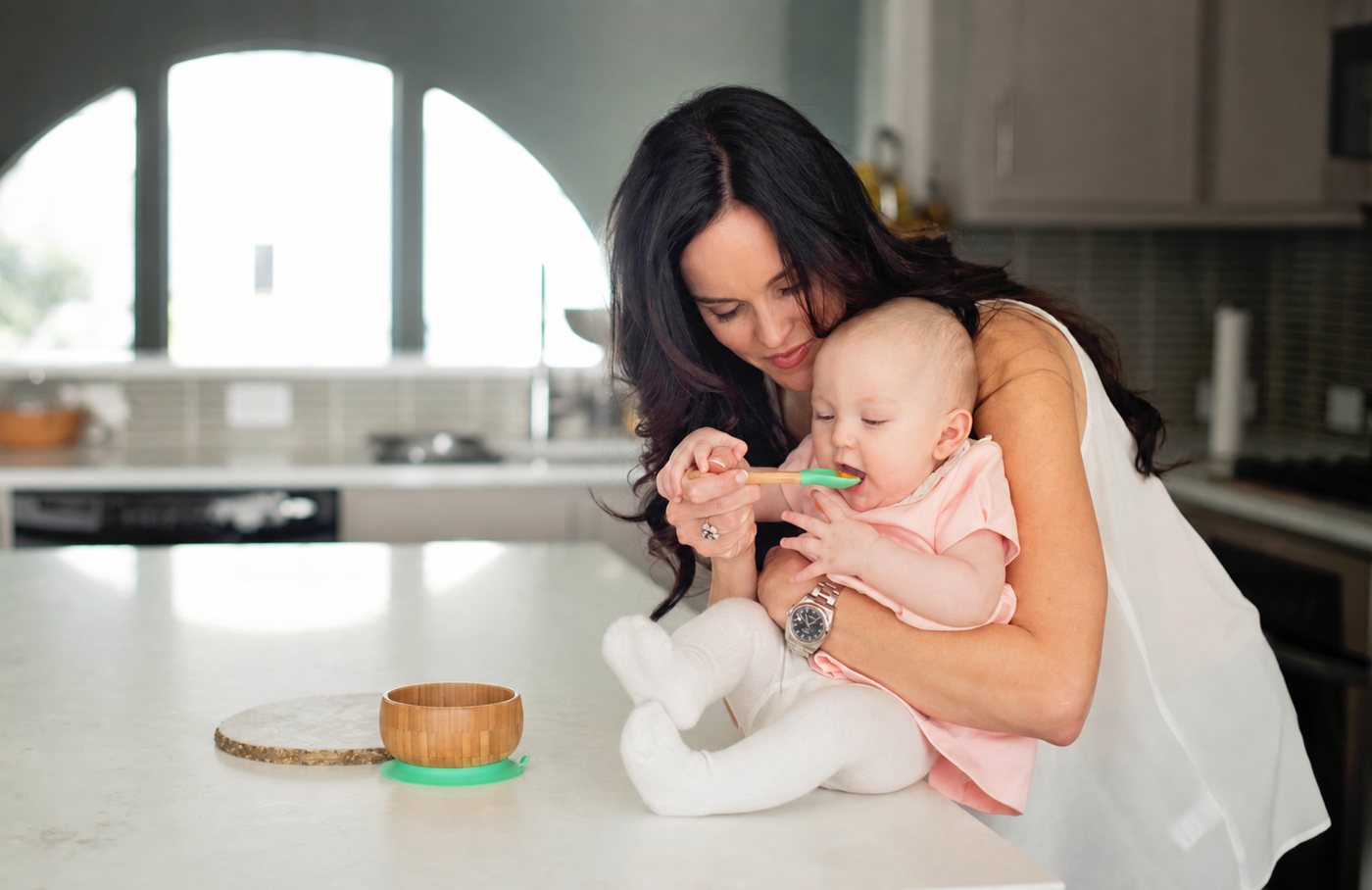 A parent feeds their baby with a bamboo-handled silicone spoon beside an Avanchy bamboo bowl with green suction.